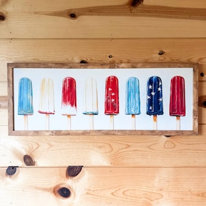 May include: A framed print showing eight popsicle illustrations in red, white, and blue. The popsicles are on wooden sticks and are arranged in a row. The artwork is in a rectangular, light brown wooden frame.