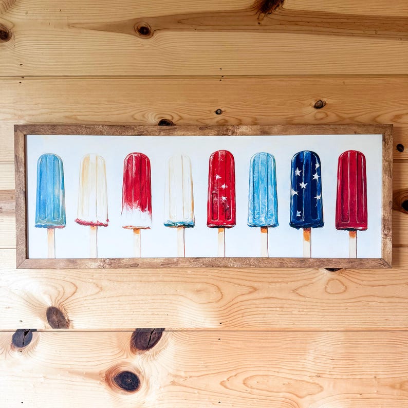 May include: A framed print showing eight popsicle illustrations in red, white, and blue. The popsicles are on wooden sticks and are arranged in a row. The artwork is in a rectangular, light brown wooden frame.