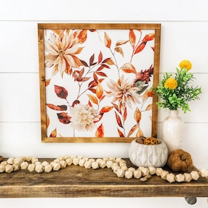 May include: A framed watercolor print of fall foliage and flowers in shades of orange, red, and white. The print is displayed on a wooden shelf with a decorative bowl and wooden beads.