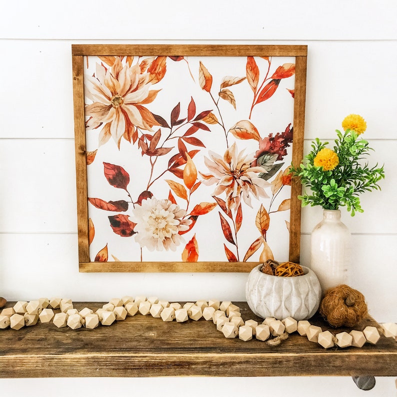 May include: A framed watercolor print of fall foliage and flowers in shades of orange, red, and white. The print is displayed on a wooden shelf with a decorative bowl and wooden beads.