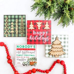 May include: Four wooden signs with a Christmas theme. The signs are decorated with red and green buffalo plaid and feature wooden cutouts of Christmas trees. The signs read "happy holidays", "Nobody's walking out on this fun old-fashioned family Christmas. We're all in this together. - Clark Griswold"