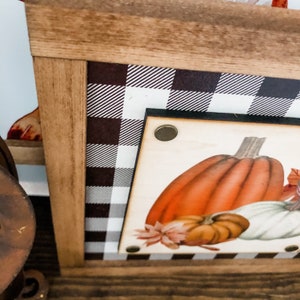 Peut inclure: Panneau en bois encadr&eacute; sur le th&egrave;me de l'automne. Le panneau pr&eacute;sente une illustration de citrouille avec des citrouilles orange, blanches et marron. Le cadre a un motif &agrave; carreaux noir et blanc et une bordure en bois naturel.