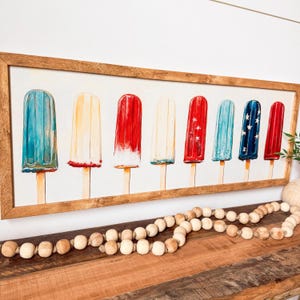 May include: A framed print with watercolour illustrations of ice lollies in red, white, and blue. The lollies are on wooden sticks and are arranged in a row. The frame is made of light brown wood, and the background is white.