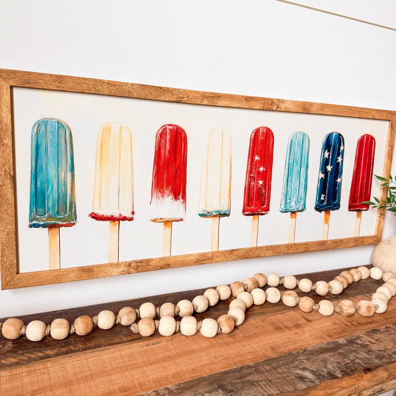 May include: A framed print with watercolour illustrations of ice lollies in red, white, and blue. The lollies are on wooden sticks and are arranged in a row. The frame is made of light brown wood, and the background is white.