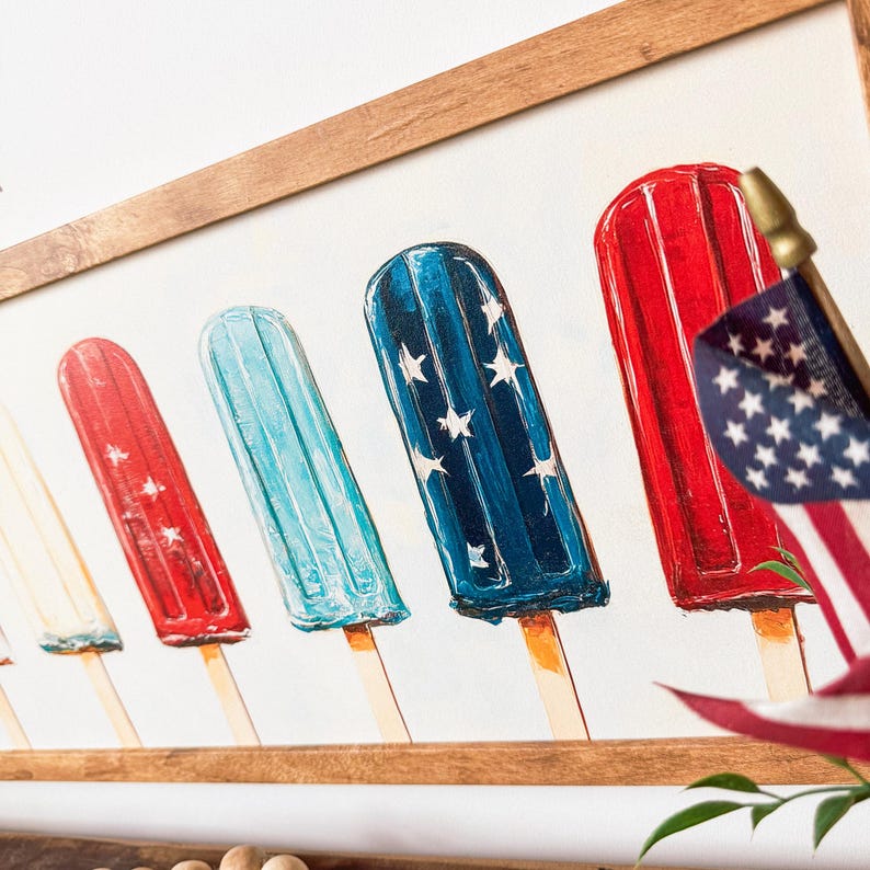 May include: A framed print depicting popsicle illustrations in red, white, and blue, with star details. The artwork has a patriotic theme, with a wooden frame. The popsicles are reminiscent of a summer treat, suitable for home decoration.