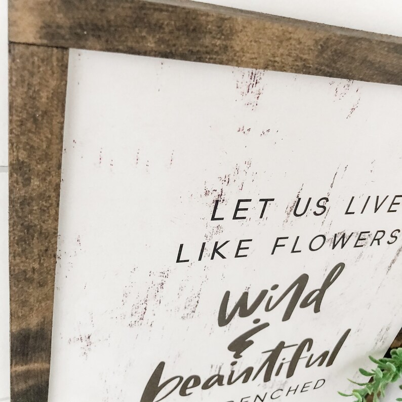 Pu&ograve; includere: Un cartello di legno bianco con vernice rovinata e lettere nere che recitano "Let us live like flowers wild & beautiful" con una piccola pianta verde nell'angolo.