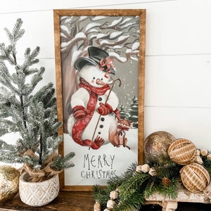 May include: A framed winter scene with a snowman holding a stick and a fox sitting beside him. The snowman is wearing a red scarf and a black top hat. The background is a snowy forest with the text "Merry Christmas" at the bottom.