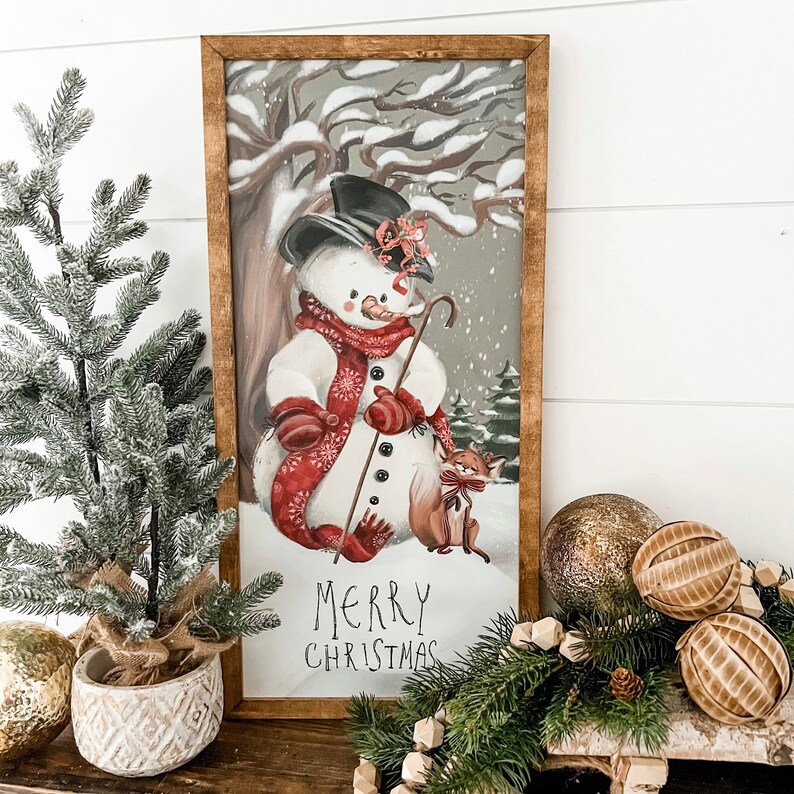 May include: A framed winter scene with a snowman holding a stick and a fox sitting beside him. The snowman is wearing a red scarf and a black top hat. The background is a snowy forest with the text "Merry Christmas" at the bottom.