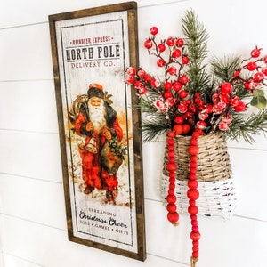 May include: A vintage-style wooden sign with a red and white illustration of Santa Claus. The sign reads "Reindeer Express North Pole Delivery Co." and "Spreading Christmas Cheer Toys Games Gifts".