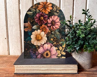 Puede incluir: Una impresión floral arqueada con un ramo de flores naranjas, rosas y crema sobre un fondo oscuro con detalles dorados. La impresión descansa sobre un libro negro grueso, con una pequeña planta en maceta a la derecha.