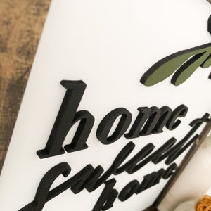 Puede incluir: Un letrero de madera blanco con letras 3D negras que dicen "home sweet home". El letrero tiene un dise&ntilde;o de hoja verde en la esquina superior derecha.