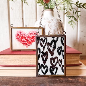 May include: Two small, rectangular glass frames with brass-colored metal edges are suspended by chains. One frame features a red heart design, while the other displays a pattern of black hearts on a white background. The frames rest on a stack of books.