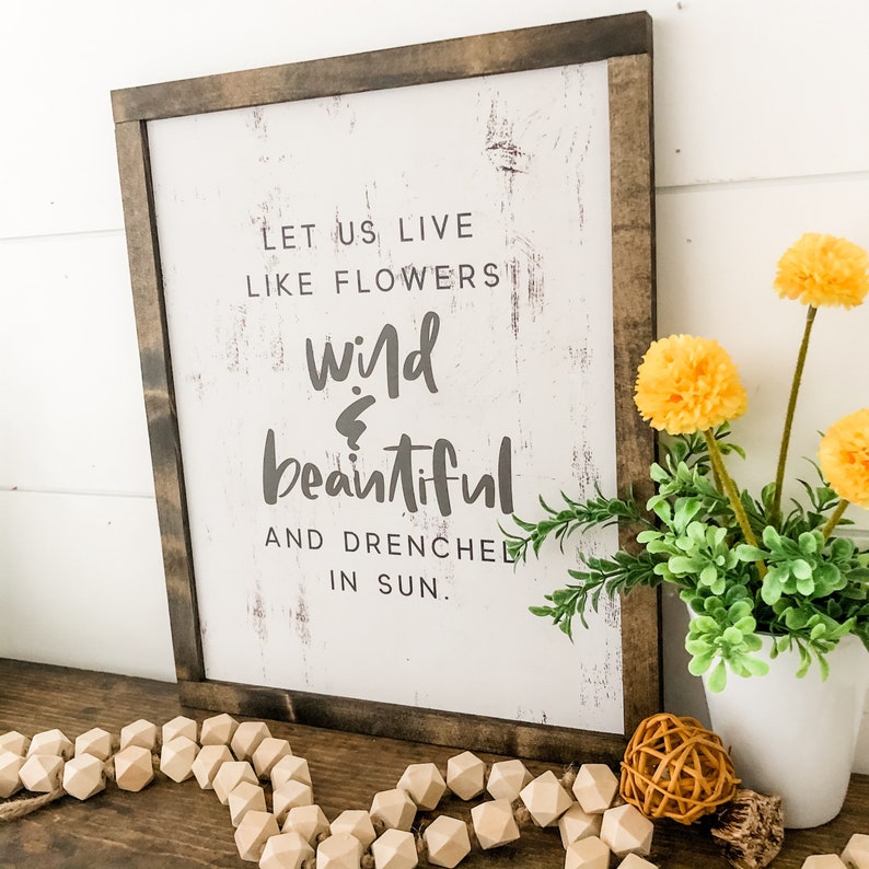 Pu&ograve; includere: Un cartello di legno bianco con una finitura usurata, con il testo "Let us live like flowers wild & beautiful and drenched in sun."