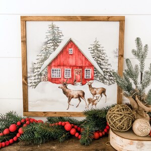 May include: A framed watercolor painting of a red cabin in a snowy forest with two deer and a fawn in the foreground. The cabin has a red door with a wreath and windows with white trim.