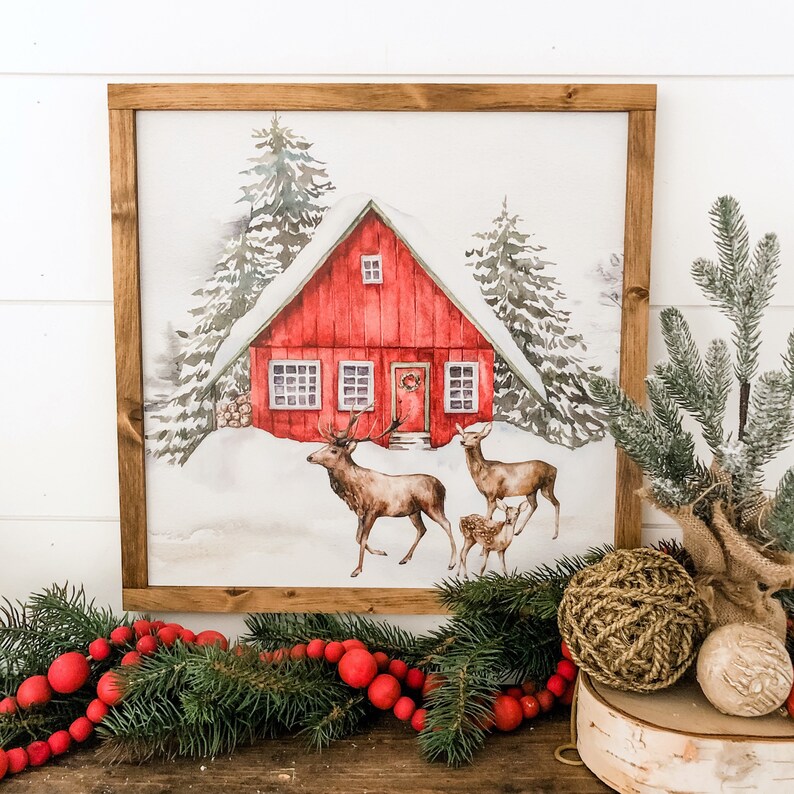May include: A framed watercolor painting of a red cabin in a snowy forest with two deer and a fawn in the foreground. The cabin has a red door with a wreath and windows with white trim.