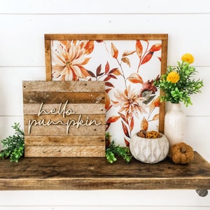 May include: A wooden sign with the text "hello pumpkin" in a rustic style, displayed on a shelf with a floral print background, a white vase with dried flowers, and a decorative bowl with dried fruit.