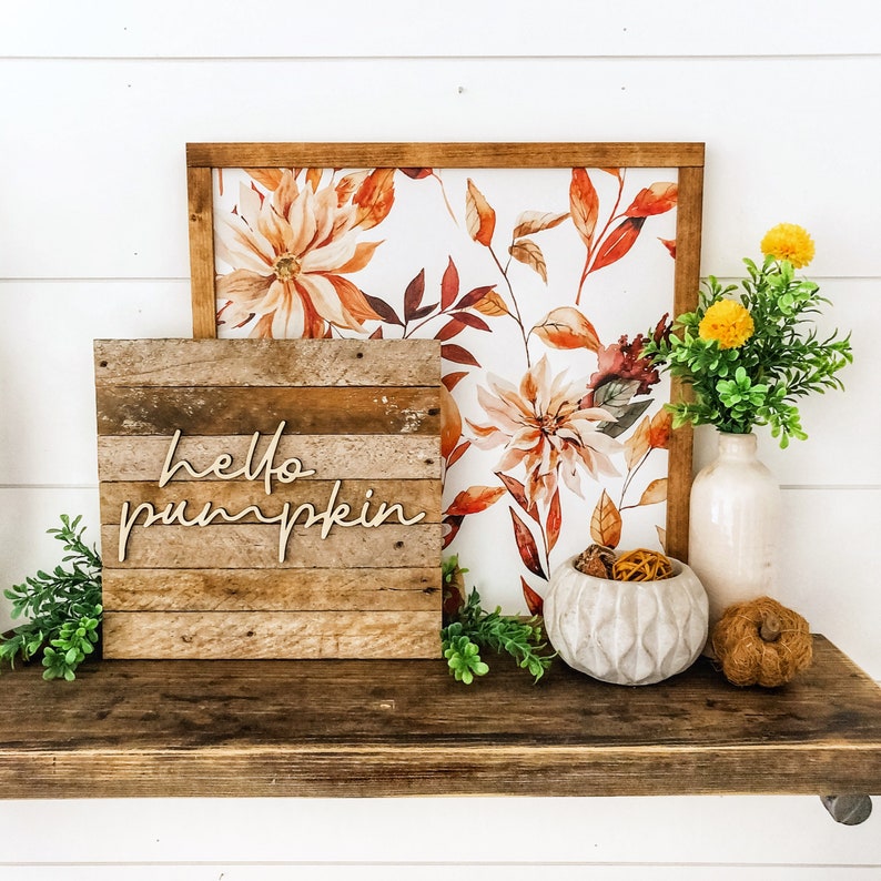 May include: A wooden sign with the text "hello pumpkin" in a rustic style, displayed on a shelf with a floral print background, a white vase with dried flowers, and a decorative bowl with dried fruit.