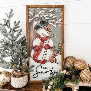 May include: A framed winter-themed illustration of a snowman wearing a red scarf and hat, holding a stick. The snowman is standing in a snowy forest with a small fox beside it. The text "Let it Snow" is written below the snowman.