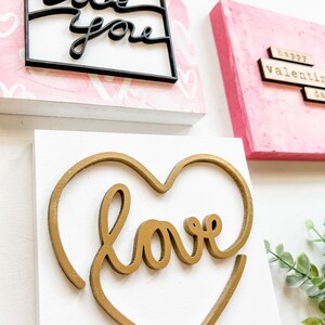 May include: Three wooden signs with Valentine's Day themes. The first sign is pink with white hearts and the words "love you" in black. The second sign is pink with the words "happy valentine's day" in brown. The third sign is white with a gold heart and the word "love" in gold.