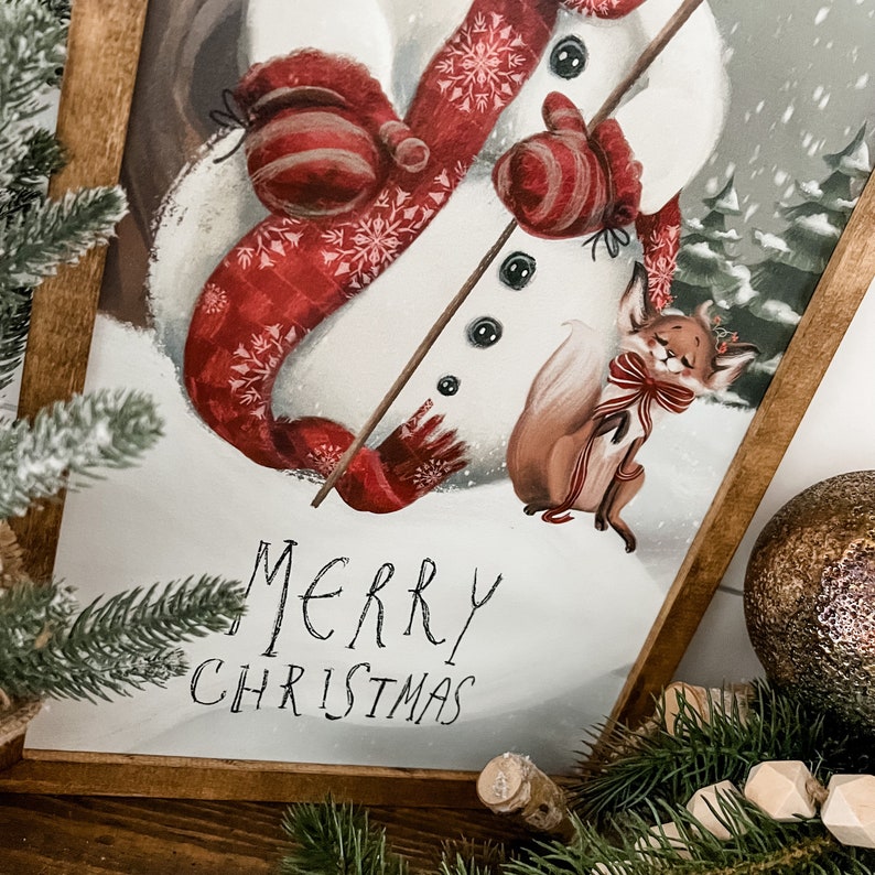 May include: A framed winter scene with a snowman wearing a red scarf and hat, holding a stick. A fox with a red bow is standing next to the snowman. The text "Merry Christmas" is written below the snowman.