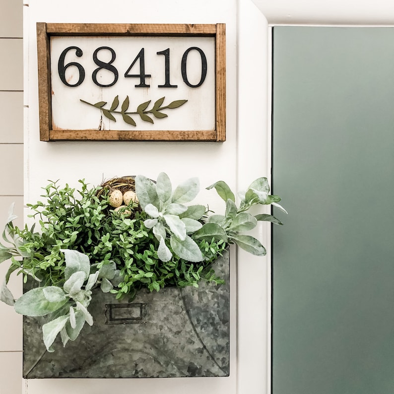 May include: A rustic wooden sign with the number "68410" in black, and a green leaf design. Below, a galvanized metal planter is filled with greenery, including lamb's ear and small green plants. A small bird's nest with eggs is nestled within.