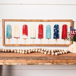 May include: A framed watercolour painting depicting seven colourful ice lollies on wooden sticks. The lollies are painted in red, white, and blue, some with star designs. The artwork is displayed on a wooden shelf.