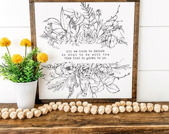 May include: A framed white sign with a dark brown wooden frame. The sign features a black and white floral design and the quote, "all we have to decide is what to do with the time that is given to us." A small white pot with yellow flowers sits to the left.