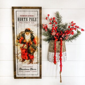 May include: A wood framed sign with a vintage illustration of Santa Claus delivering gifts. The sign reads "Reindeer Express North Pole Delivery Co. Spreading Christmas Cheer Toys Games Gifts". A red berry sprig is in a white and brown wicker basket.