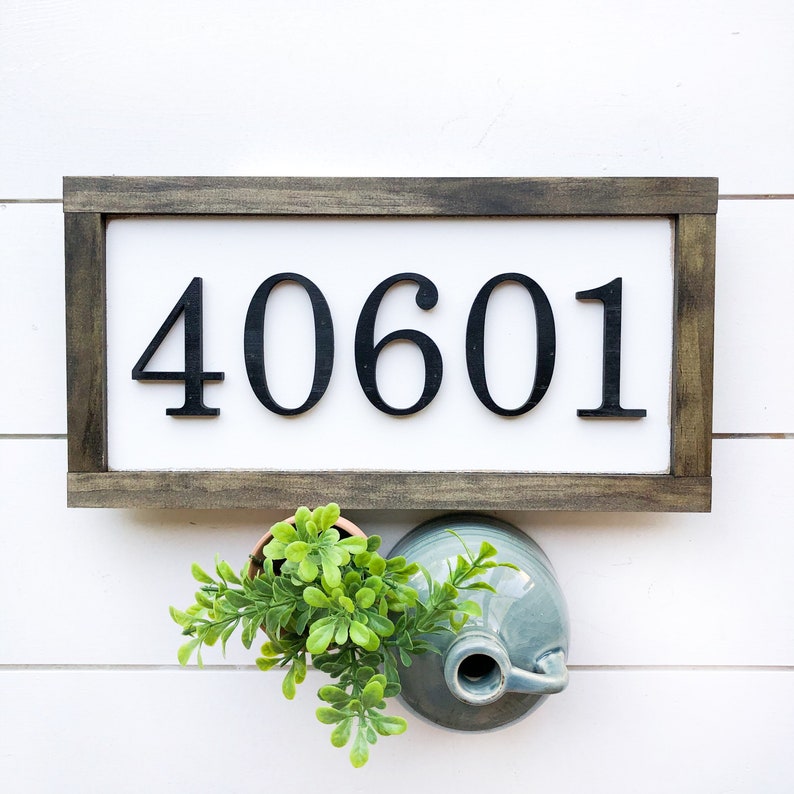 May include: A rectangular wooden sign with the numbers "40601" in black, set against a white background. The frame is a dark brown color. Below the sign are a small potted plant and a blue ceramic jug.