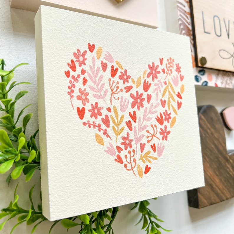 May include: A white wooden sign with a pink, red, and yellow floral heart design. The heart is made up of small flowers, leaves, and hearts.