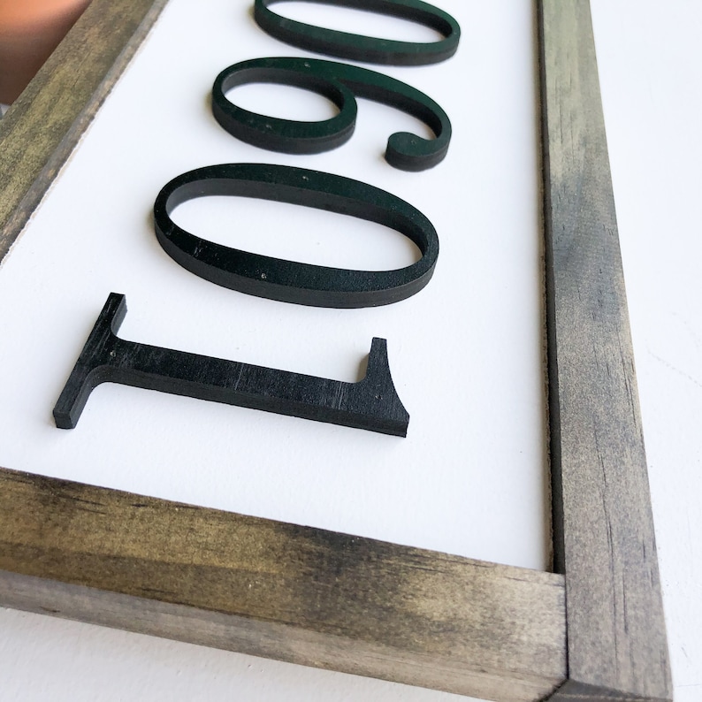 May include: A rectangular wooden sign with the numbers 1060 in black, three-dimensional lettering. The numbers are set against a white background and framed by a weathered gray and brown wooden border. The sign is likely for home decor.