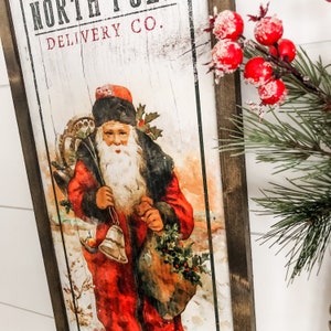 May include: A vintage-style wooden sign with a distressed white background. The sign features the text "North Pole Delivery Co." and a depiction of Santa Claus carrying a bag of gifts and a bell. The sign is framed in dark wood.
