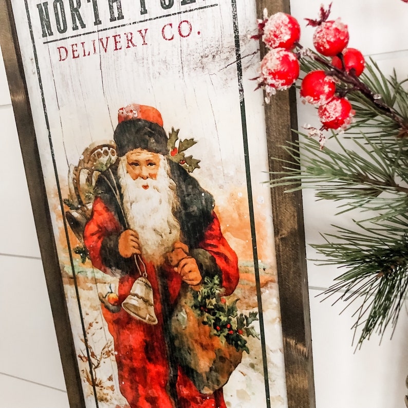May include: A vintage-style wooden sign with a distressed white background. The sign features the text "North Pole Delivery Co." and a depiction of Santa Claus carrying a bag of gifts and a bell. The sign is framed in dark wood.
