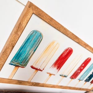 May include: A framed print depicting a row of colourful ice lollies on wooden sticks. The ice lollies are painted in shades of red, white, and blue, with a light brown wooden frame. The artwork evokes a summer theme.