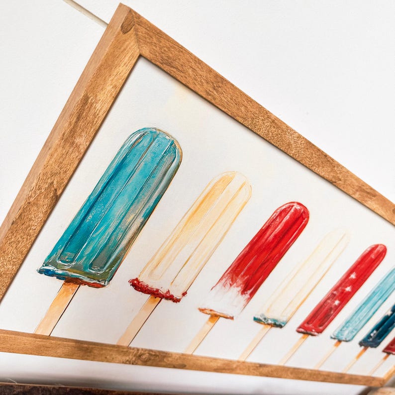 May include: A framed print depicting a row of colourful ice lollies on wooden sticks. The ice lollies are painted in shades of red, white, and blue, with a light brown wooden frame. The artwork evokes a summer theme.