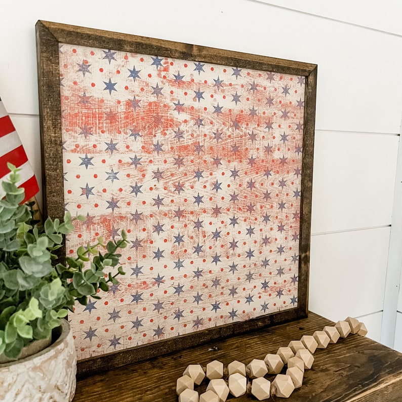 May include: A framed wood sign with a distressed American flag design featuring red, white, and blue stars and dots. The sign is perfect for patriotic decor.