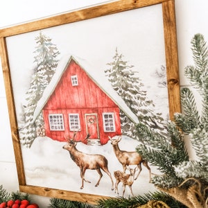 May include: A watercolor painting of a red cabin in a snowy forest with three deer. The cabin has a wreath on the door and snow on the roof. The deer are brown and white and are standing in the snow.
