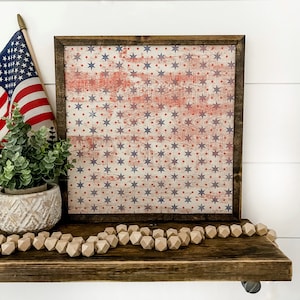 May include: A framed wood sign with a distressed American flag design featuring red, white, and blue stars and dots. The sign is displayed on a wooden shelf with a beige wooden bead garland.
