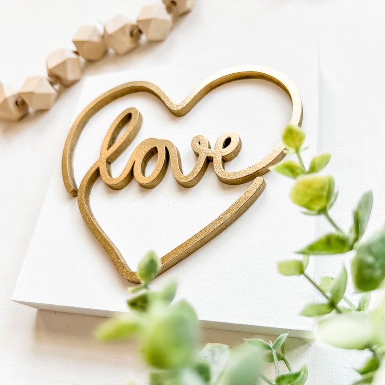 May include: A white wooden sign with a gold heart cutout and the word "love" written in a cursive font inside the heart.