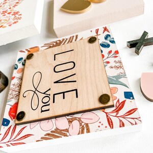 May include: A wooden sign with a floral pattern and the words "Love you" in black lettering. The sign has a light wood background and is decorated with gold accents.