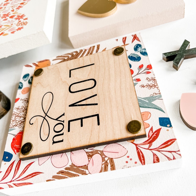 May include: A wooden sign with a floral pattern and the words "Love you" in black lettering. The sign has a light wood background and is decorated with gold accents.
