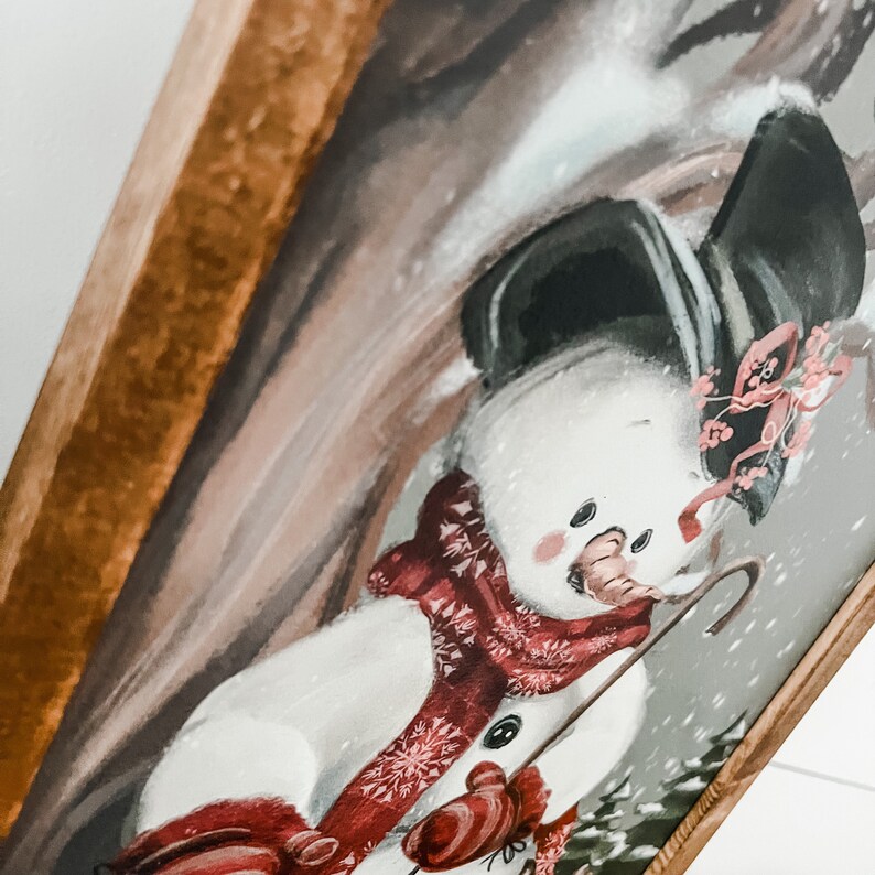 May include: A framed painting of a snowman wearing a red and white scarf and a black hat with a red flower. The snowman is holding a brown stick and is standing in front of a brown and white background.