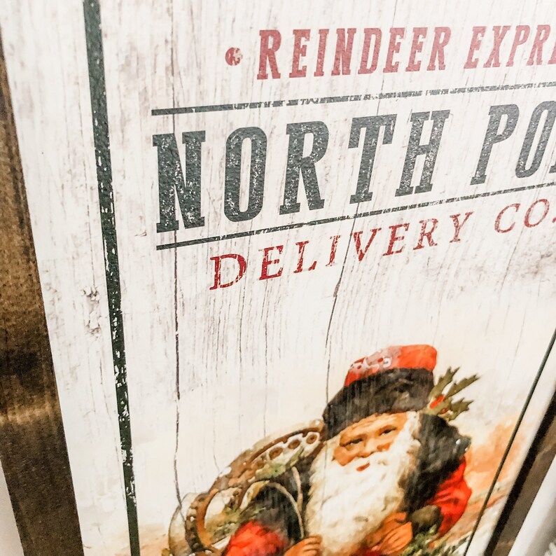 May include: A vintage-style sign with the text "Reindeer Express North Pole Delivery Co." in red and black lettering on a white wood background. A cartoon image of Santa Claus in a red and black suit is in the bottom right corner of the sign.
