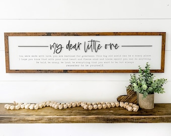 May include: A wooden framed sign with the text "my dear little one" in a cursive font. Below the title is a printed message. A wooden bead garland and a small potted plant sit on a wooden shelf below the sign.