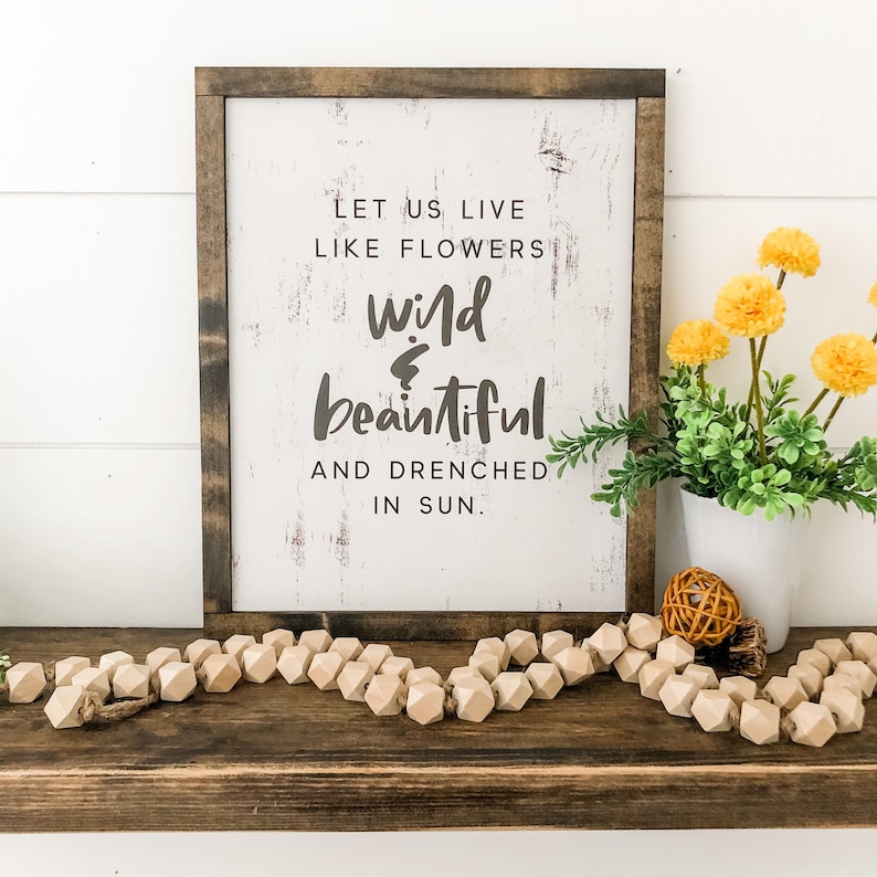 Pu&ograve; includere: Un cartello di legno con vernice bianca e un aspetto usurato. Il cartello recita "Let us live like flowers wild & beautiful and drenched in sun."