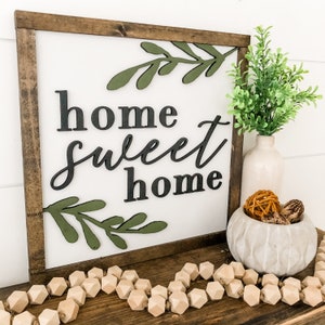 Puede incluir: Un letrero de madera con las palabras "home sweet home" en letras negras y ramas de hojas verdes. El letrero est&aacute; en un estante de madera con una peque&ntilde;a maceta de hormig&oacute;n con una esfera marr&oacute;n y una planta verde.