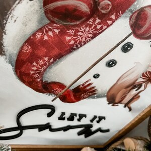 May include: A white snowman with a red and white patterned scarf and a black stick for arms. The snowman has black eyes and a black button nose. The background is white with the words "Let it Snow" in black.