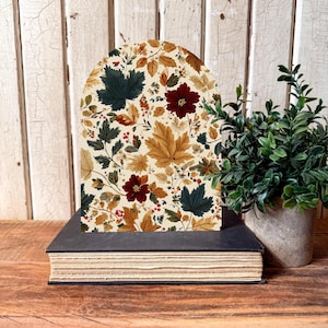 May include: A decorative arched panel featuring a fall-themed floral pattern with maple leaves, berries, and flowers in shades of burgundy, gold, and green. The panel rests on a dark book, with a small potted plant to the right.