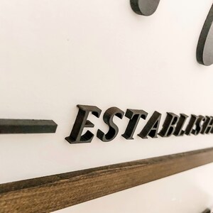 May include: Close-up of a black wooden sign with the word "ESTABLISHED" in raised letters. The sign is mounted on a white wall with a dark wood frame.