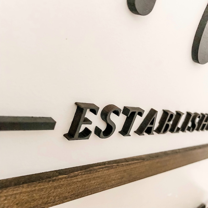 May include: Close-up of a black wooden sign with the word "ESTABLISHED" in raised letters. The sign is mounted on a white wall with a dark wood frame.